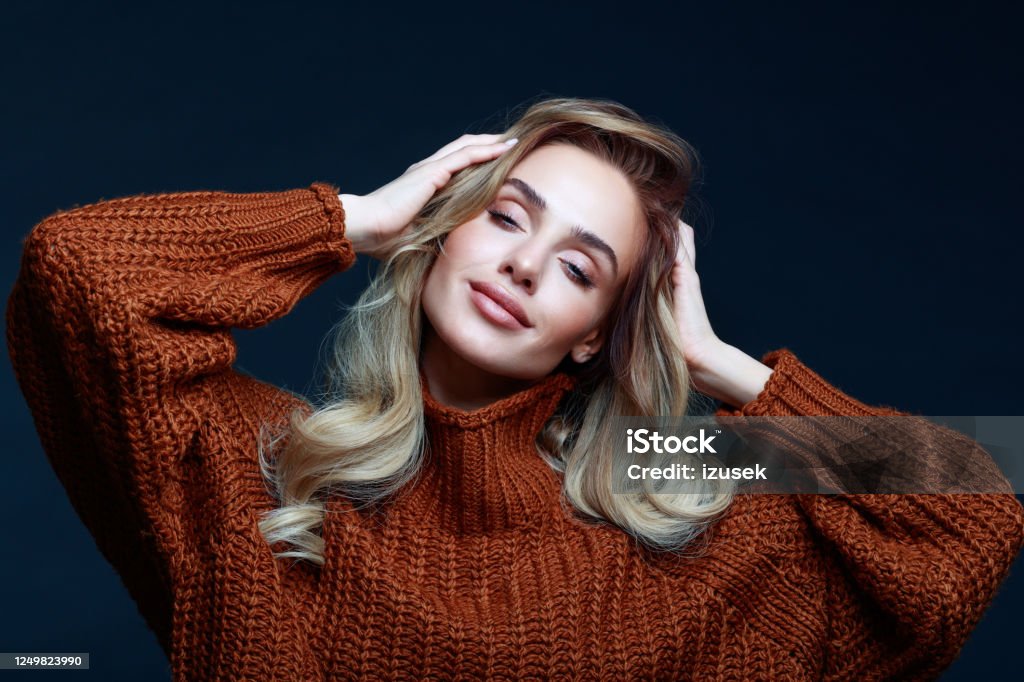 Autumn portrait of blond hair woman in brown sweater Fashion portrait of long hair blond young woman wearing brown sweater, smiling with eyes closed and raised hands. Studio shot against black background. Autumn Stock Photo