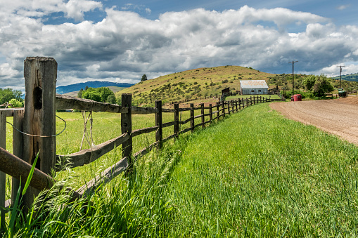 Long Split Rail Fence along a grassy field near a dirt roat leading up to a metal barn under the cloudy sky