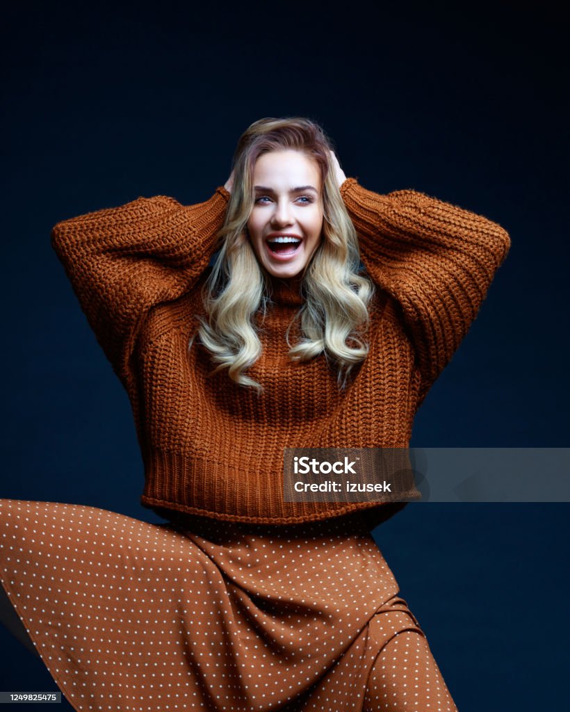 Autumn portrait of excited woman in brown clothes Fashion portrait of long hair blond young woman wearing brown sweater and skirt, laughing with raised hands. Studio shot against black background. Women Stock Photo