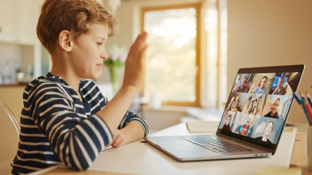 istock Cute Little Boy Uses Laptop with Conference Video Call Software to Talk with Group of Relatives and Friends. He Greets His Family Member and They Wave Back. Happy Family Connected Online 1249825729