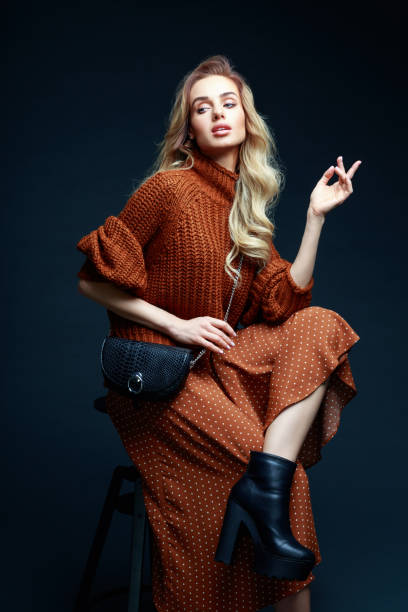 Fashion portrait of elegant woman in brown clothes, dark background Portrait of long hair blond young woman wearing brown sweater and skirt, holding black purse, sitting on chair, looking away. Studio shot against black background. upper class photos stock pictures, royalty-free photos & images
