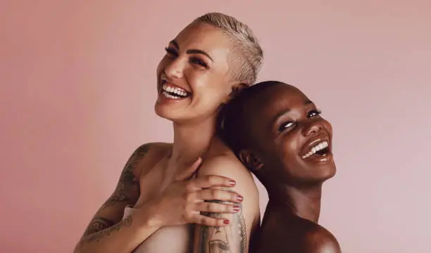 Two women with buzz cut hairstyle standing back to back and smiling at camera. Female models with beautiful skin smiling together over beige background.