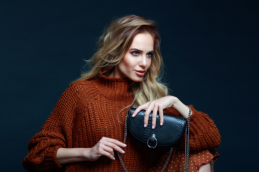 Portrait of long hair blond young woman wearing brown sweater and skirt, holding black purse, looking away. Studio shot against black background.