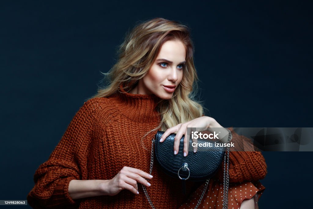 Fashion portrait of elegant woman in brown sweater, dark background Portrait of long hair blond young woman wearing brown sweater and skirt, holding black purse, looking away. Studio shot against black background. Purse Stock Photo