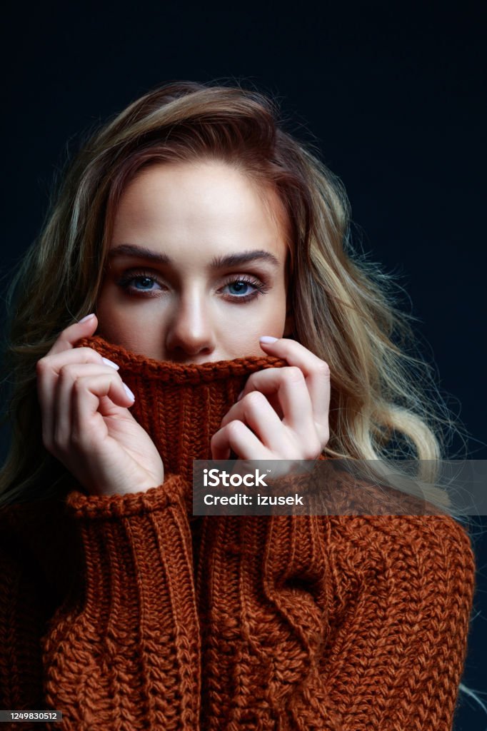Headshot of blond hair woman in brown sweater Fashion portrait of long hair blond young woman wearing brown sweater, looking at camera and covering mouth. Studio shot against black background. Sweater Stock Photo