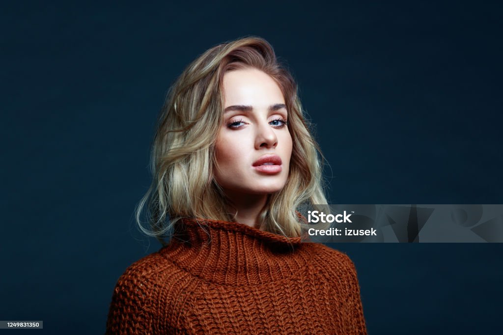 Headshot of blond hair woman in brown sweater Fashion portrait of long hair blond young woman wearing brown sweater, looking at camera. Studio shot against black background. Blond Hair Stock Photo