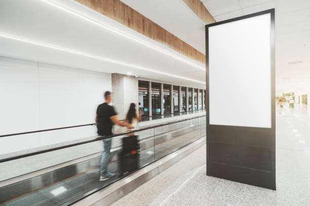 Vertical banner mockup in an airport An indoor arrival or departure area of a modern airport with blurry silhouettes of  passengers passing by on travelator and a vertical empty mockup of an information LCD panel or an advert blillboard airport travelator stock pictures, royalty-free photos & images