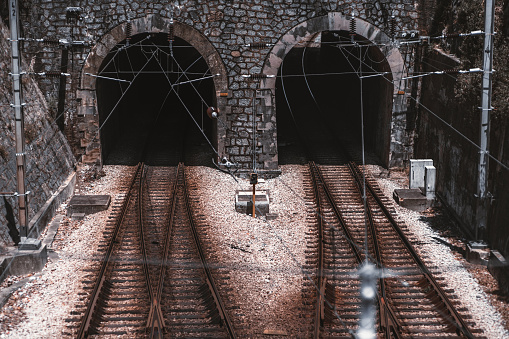 Two entrances to dark railway tunnels of arc shape and stone wall, with railroad junctions in front and power cables above and on the sides, pebble on the ground, sunny day, Sintra, Portugal