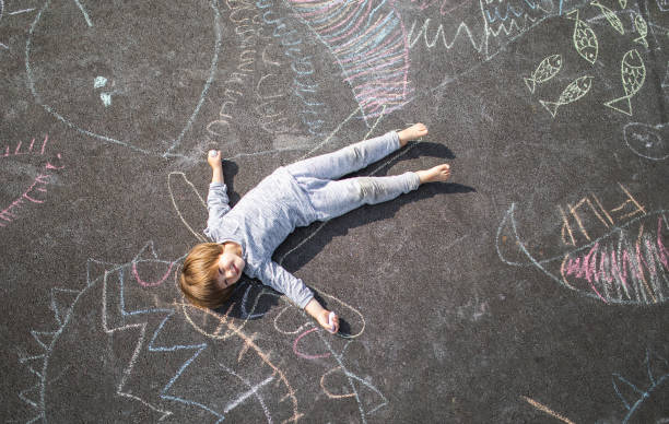 young boy lying down on the asphlat doodled with chalk - child chalking imagens e fotografias de stock