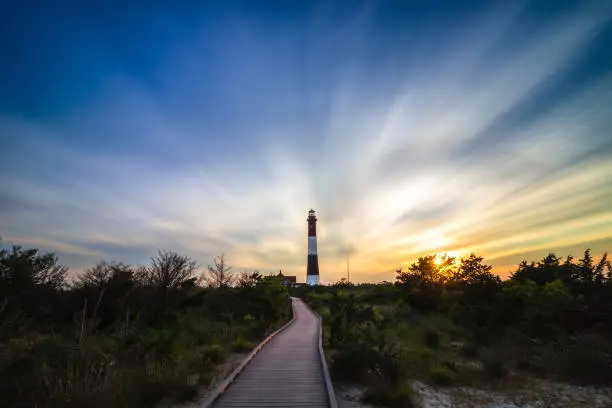 Photo of Wooden boardwalk leading to a tall stone lighthouse, as sunset clouds streak across the sky overhead. Fire Island NY