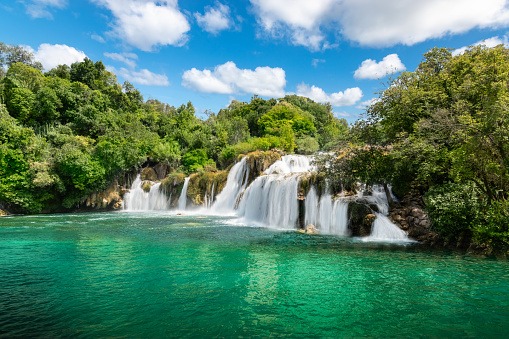Breathtaking  panoramic landscape with flowing waterfall, Buk Skradinski, in the National Park of Croatia. Colorful tourist summer vacation  highlight without people. Bright and wide image with blue sky, white clouds, forest trees, green lake water  and misty, dreamy waterfall texture.