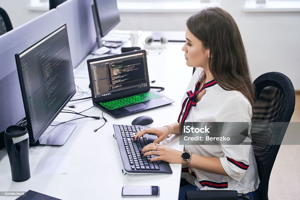 Beautiful female junior software developer working on computer in IT office, sitting at desk and coding, working on a project in software development company or technology startup. High quality image. Beautiful young woman working on computer in IT office, sitting at desk and coding, working on a project in software development company or technology startup. Computer Programmer Stock Photo
