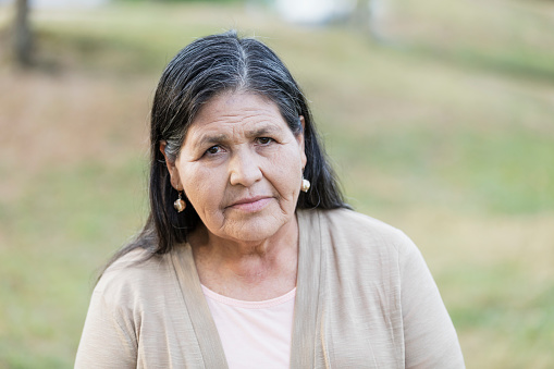 A senior Hispanic woman in her 60s standing outdoors in a park, looking at the camera with a serious expression on her face.