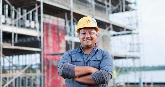 A young Hispanic man in his 20s working at a construction site, wearing a hardhat and safety goggles. He is looking at the camera, arms crossed, smiling. A building under construction is out of focus behind him.