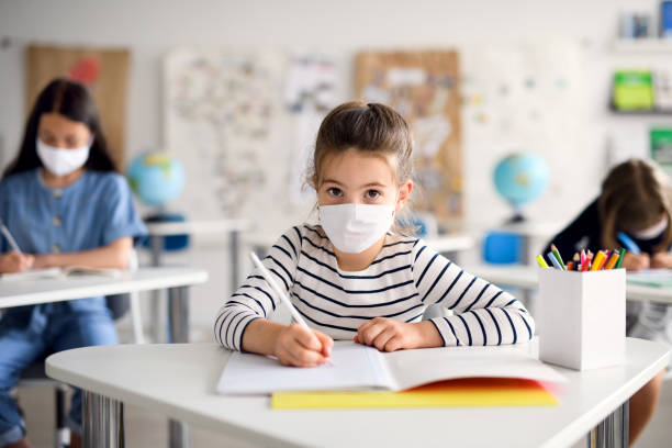Child with face mask back at school after covid-19 quarantine and lockdown, writing. Front view portrait of child with face mask back at school after covid-19 quarantine and lockdown, writing. lockdown viewpoint photos stock pictures, royalty-free photos & images