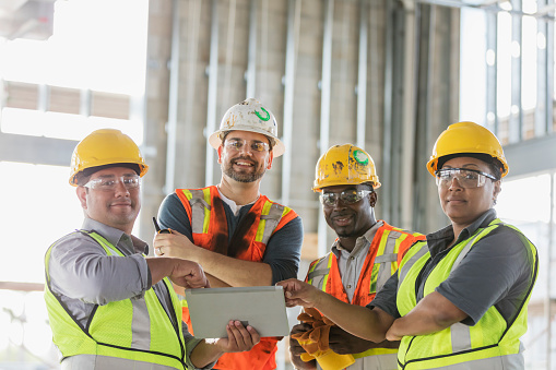 A multi-ethnic group of four construction workers wearing hardhats and safety vests, having a meeting inside the structure being built. A young Hispanic man is holding a digital tablet and they are all smiling and looking at the camera. The worker on the right is a mature African-American woman in her 40s.