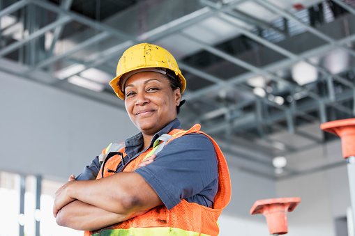 A mature African-American woman in her 40s wearing a hardhat and reflective vest, a construction worker or building contractor standing with her arms crossed, smiling at the camera.