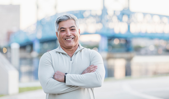 Portrait of a senior Hispanic man in his 60s standing outdoors, smiling at the camera with his arms crossed. He is on a city waterfront, bridge out of focus in the background.
