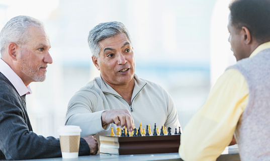 A group of three middle-aged and senior multi-ethnic men in their 50s and 60s playing a game of chess. The view if from over the shoulder of the African-American man. The senior Hispanic man is reaching for a piece to make his move.
