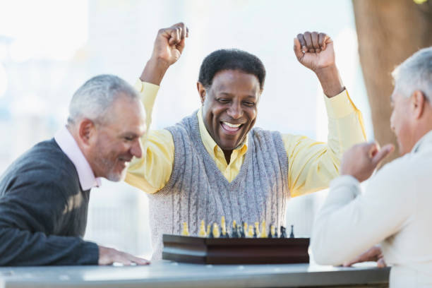 Men playing chess, winning move A group of three middle-aged and senior multi-ethnic men in their 50s and 60s playing a game of chess. The view if from over the shoulder of the senior Hispanic man. His African-American opponent is laughing, fists in the air, declaring victory. senior chess stock pictures, royalty-free photos & images