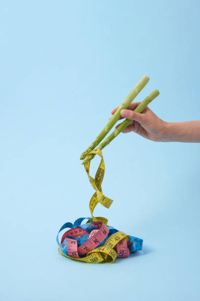 Woman's hand is taking measure tapes by asparagus spears as sticks. Woman's hand with asparagus sticks is taking measure tapes as a colorful noodle against pastel blue background, copy space. Colored measuring tapes as Japanese or Chinese food. eating asparagus stock pictures, royalty-free photos & images