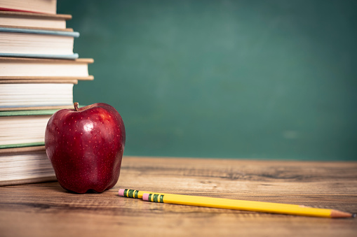 Back to school.  Red apple, pencil on top of a stack of textbooks on wooden school desk in front of green chalkboard.  Classroom setting.