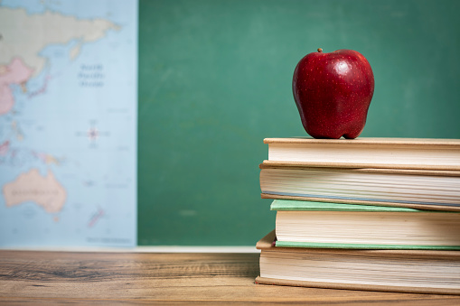 Back to school.  Red apple on top of a stack of textbooks on wooden school desk in front of green chalkboard and map.  Classroom setting.