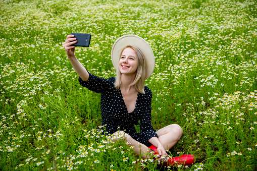 Blonde woman in black dress sitting in countryside chamomiles flowers field and using mobile phone