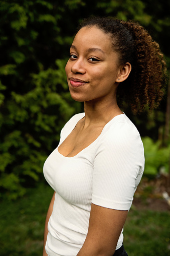 Portrait of beautiful mixed-race teenage girl backyard in nature. She is standing on the grass, looking at the camera with a smile. Vertical outdoors waist shot with copy space.