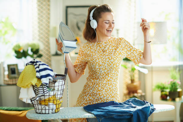 woman ironing on ironing board while listening to the music happy modern middle age woman in yellow dress with washed clothes basket ironing on ironing board while listening to the music with headphones at modern home in sunny day. iron appliance stock pictures, royalty-free photos & images