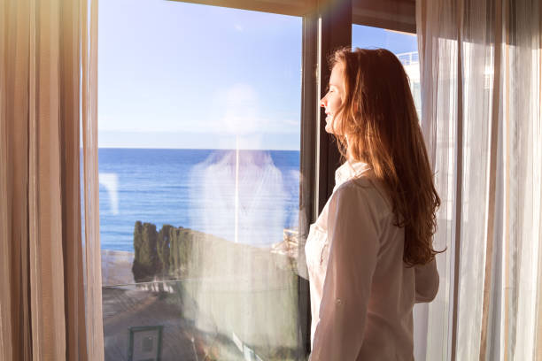 istock Lovely long-haired woman in a white shirt standing inside by a panoramic window overlooking the sea and enjoying the sun with her eyes closed. 1249886774