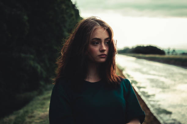 Young day dreaming, pensive and sad looking young woman standing alone outdoors close to river in sunset twilight. Thinking, looking to the horizon. Real People Youth Culture Lifestyle Portrait.