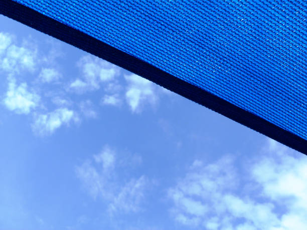 Loosely weaved breathing blue fabric sun shade material spanning over terrace or patio area. blue sky and white clouds. abstract low angle view. metal connector and chain. summer and outdoors concept.