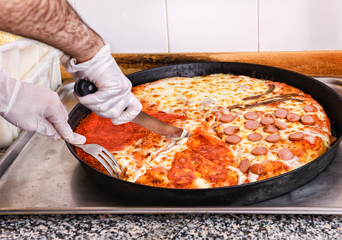 Chef slicing an Italian pizza with four assorted toppings using a knife and fork in a pizzeria kitchen in a close up on his hands