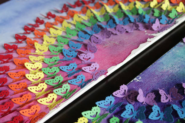 Stock photo of close-up of gay themed homemade crafting art work, using rainbow butterfly punch with cut out card and paper butterflies, embossed and glued on abstract watercolour painting that has been painted on a stretched canvas. This original craft art work of rainbow coloured made of multicoloured cut out butterfly shapes (Monarch paper butterflies - purple, blue, green, orange, yellow and red colours) is like the rainbow LGBTQI gay flag, with the colourful arts and crafts project displayed on wall as background wallpaper. imagen de primer plano de la obra de arte artesanal casero de temática gay, utilizando el ponche de mariposa arco iris con tarjeta cortada y mariposas de papel, en relieve y pegado en pintura abstracta de acuarela pintada sobre un lienzo estirado. artesan