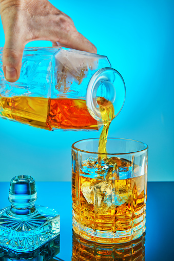 Pouring square crystal decanter with scotch tape whiskey or brandy in a crystal round glass with ice on a blue gradient background with reflection. Copy space
