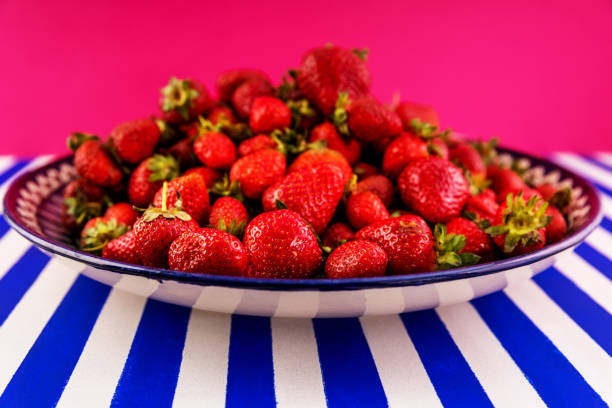 Delicious fresh strawberries on a huge earthenware dish on a blue-and-white striped table. Side view on a lilac background.