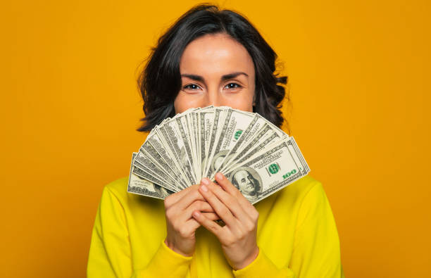 ¡aquí está mi salario! foto de primer plano de una joven con sudadera amarilla, sonriendo con los ojos, escondiendo su rostro detrás de una gran cantidad de dinero en sus manos. - agarrar fotografías e imágenes de stock