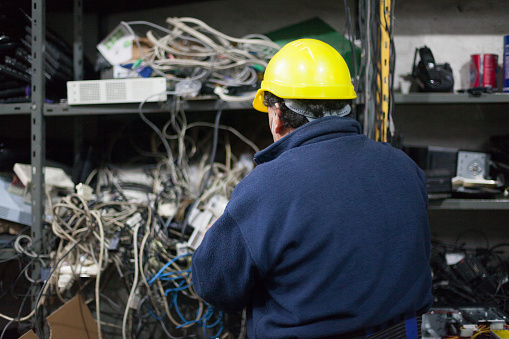 Middle aged technician, wearing protective mask and helmet, is sorting computer cords in a shelf