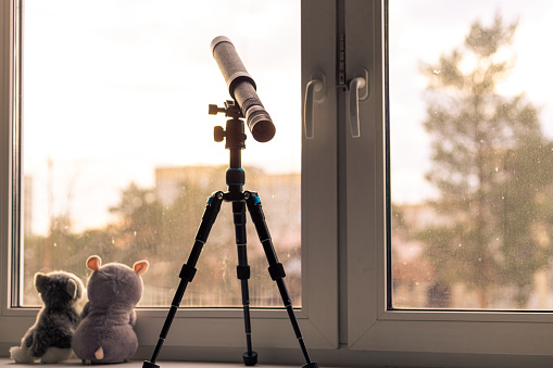 on the window sill stands on a tripod a telescope pointing at the sky, sitting next to two soft toys turned to the street, brown tinted, retro, self-isolation mode, coronavirus quarantine