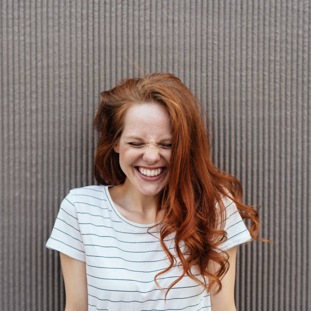 Funny portrait of a happy young redhead woman with long curled hair wearing a striped summer T-shirt while laughing with closed eyes
