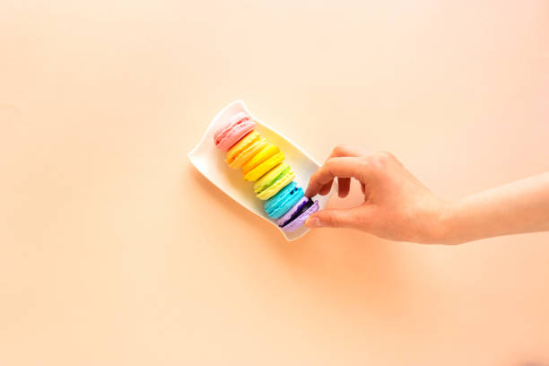 istock Minimalistic composition with six handmade macaroons of different colors lying on white rectangle plate in the right part of the photo. Cookies of pink, blue, light green, yellow, purple, and orange colors. The hand in the frame reaches for macaroons 1249967935