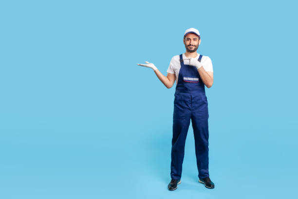istock Full length cheerful worker in blue overalls, cap and protective gloves standing, pointing at empty space 1249972846