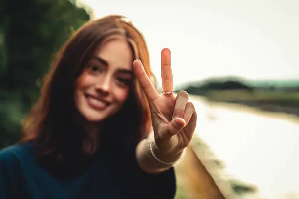 Young happy smiling woman with Peace Hand Sign - Victory Hand Sign outdoors close to a river in moody sunset twilight. Selective Focus on Peace Hand Sign.
