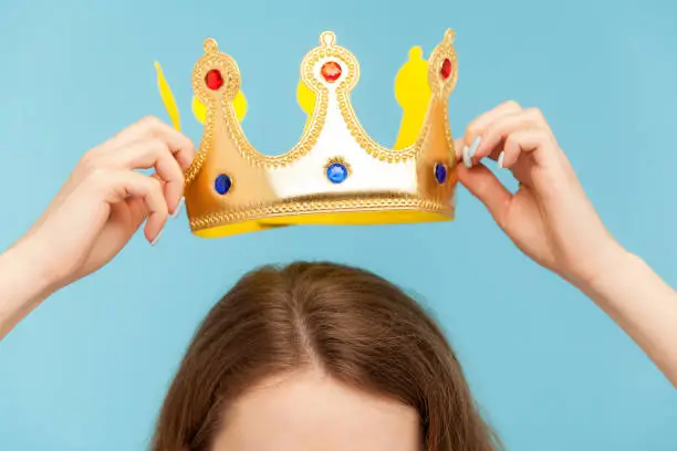 Closeup of woman putting on head golden crown, concept of awards ceremony, privileged status, superior position, self-motivation and big ambitions to achieve success. indoor studio shot, isolated