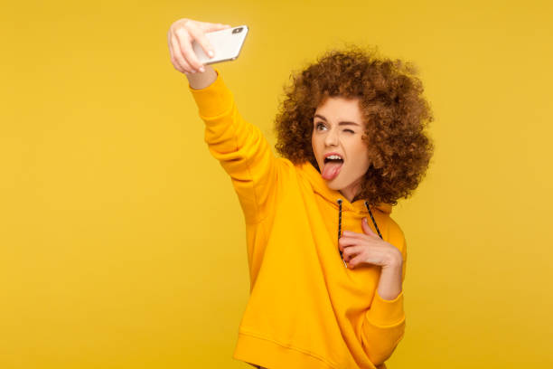 Portrait of carefree trendy curly-haired girl in urban style hoodie taking selfie and grimacing with tongue out Portrait of carefree trendy curly-haired girl in urban style hoodie taking selfie and grimacing with tongue out, having fun while doing photo, vlog broadcast. studio shot isolated on yellow background grimacing photos stock pictures, royalty-free photos & images