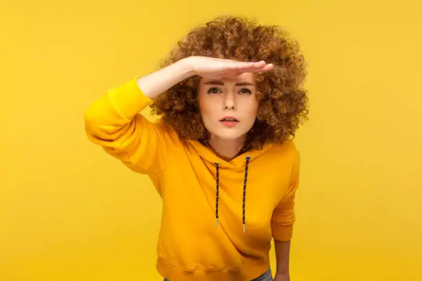 Portrait of curious curly-haired woman in urban style hoodie holding hand above eyes and peering into distance, looking far away, expecting and searching someone on horizon. studio shot isolated