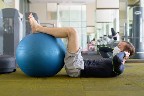 tiro de cuerpo completo de joven con máscara haciendo sentarse con pelota de ejercicio en el gimnasio durante el virus corona covid-19 - sit ups fotografías e imágenes de stock