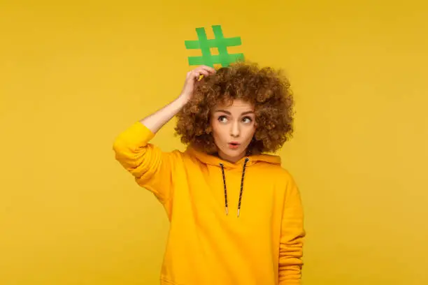 Photo of Popular blog posts, trendy content. Portrait of surprised curly-haired woman in urban style hoodie holding hashtag