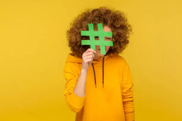 Let's look at internet trends, viral blog post. Portrait of curly-haired woman in urban style hoodie looking through hashtag sign with curious nosy expression. indoor studio shot, yellow background